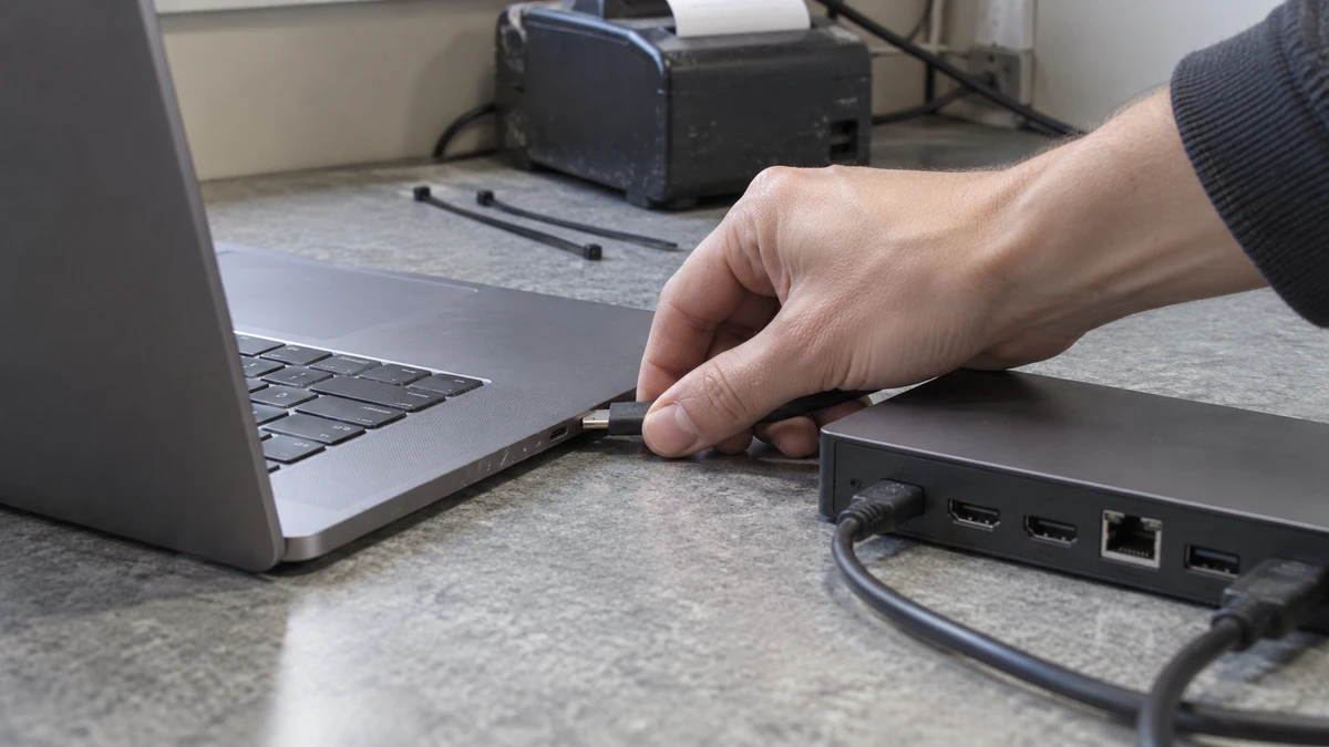 A hand connects a laptop to a dock beside a receipt printer.