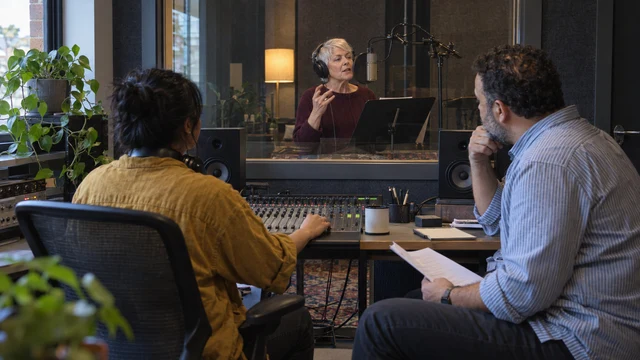 A producer and an account lead watch through the studio glass as a voice artist records a script.
