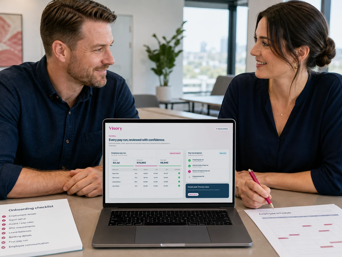 Two colleagues review payroll paperwork on a laptop in a services office.