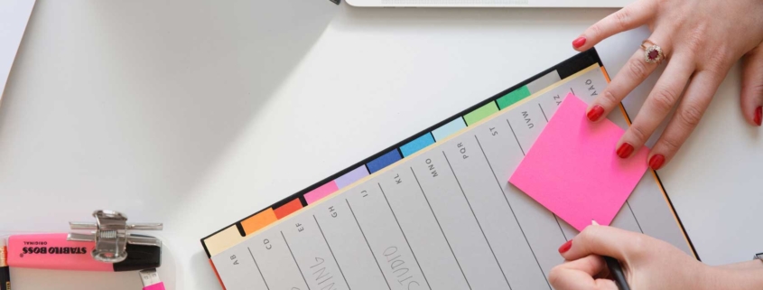 A woman prepare to write on a pink post-it note while at her desk.