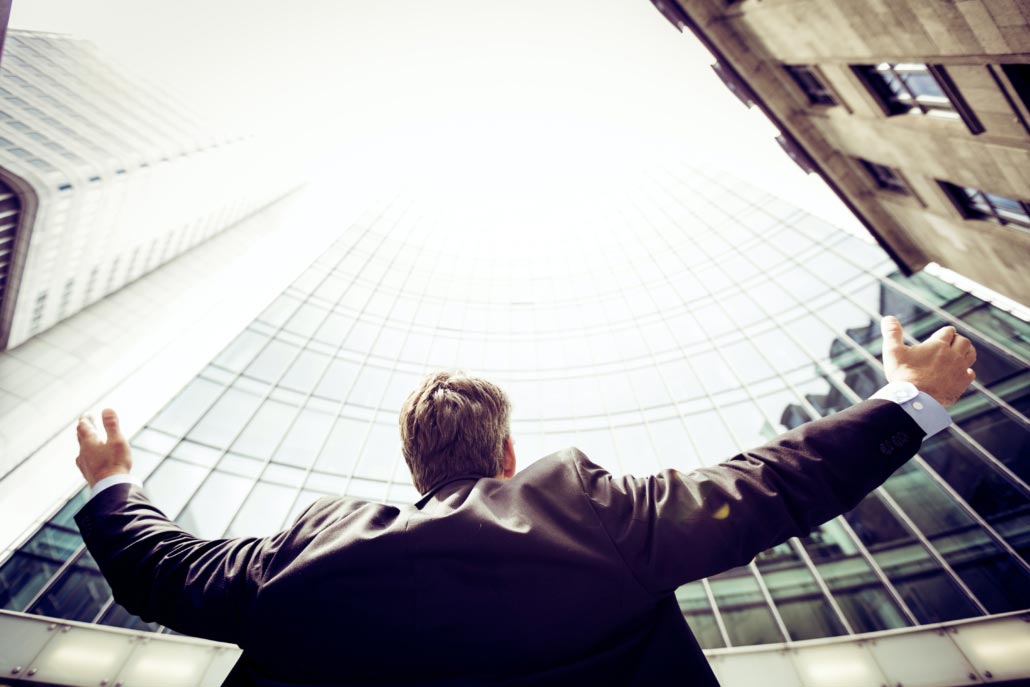 A business man looks up at a glass skyscraper with his arms raised high